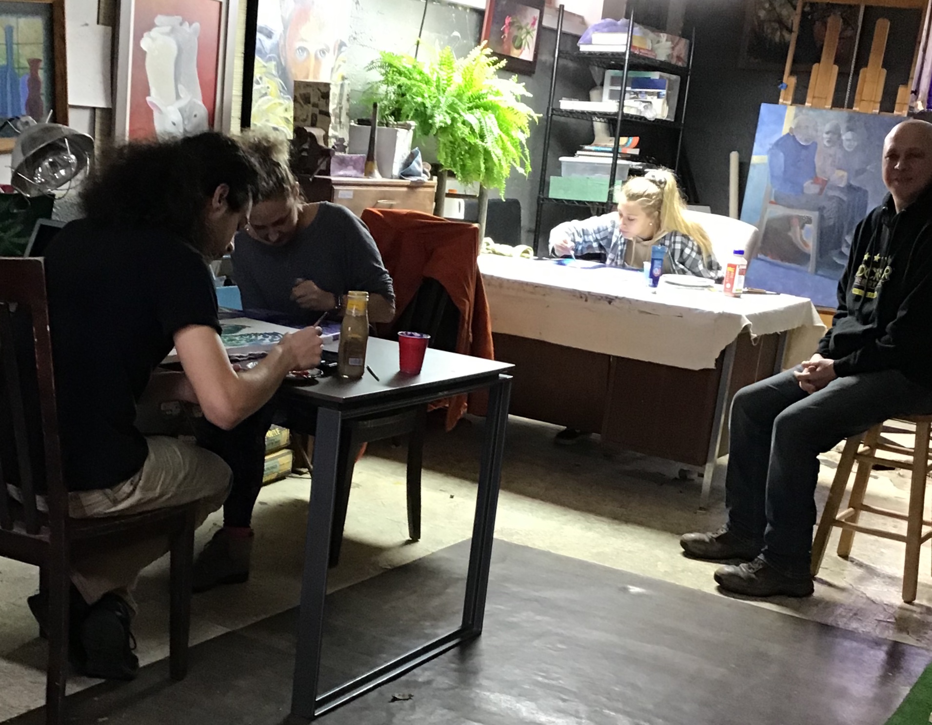People sitting at tables in an art studio, with paintings and supplies visible in the background.