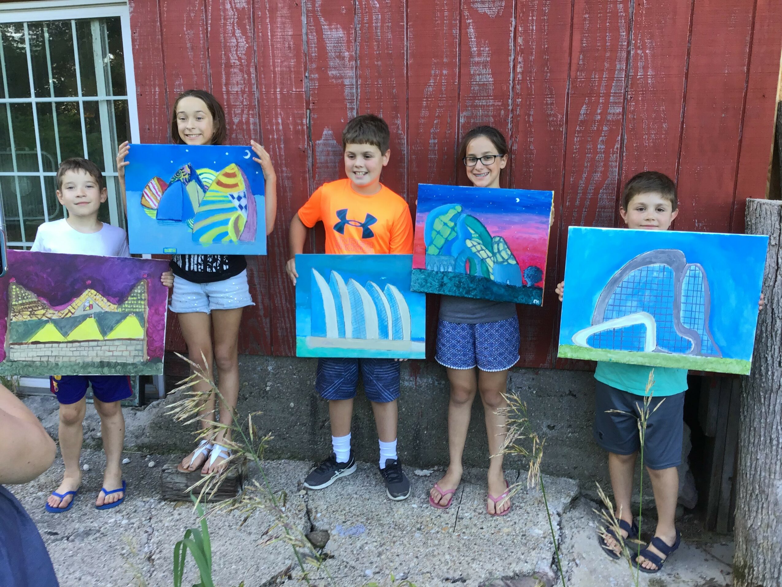 Four children and one adult holding colorful paintings outdoors against a red wooden wall, with grass and a fence nearby.