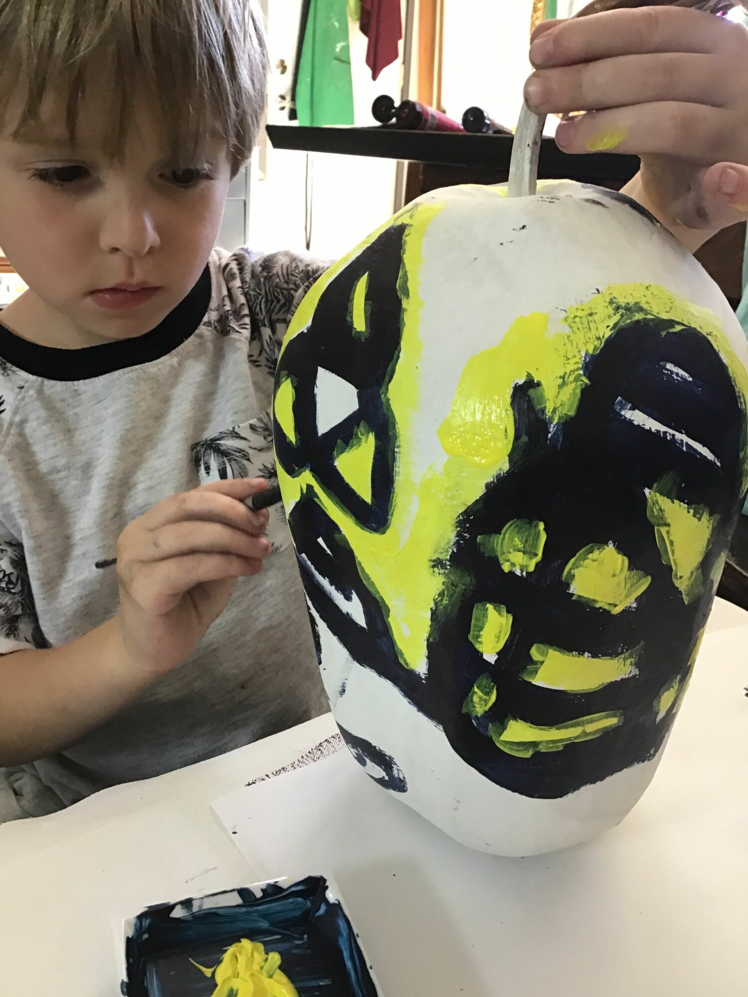 Child painting a large white object with black and yellow designs, sitting at a table with art supplies.