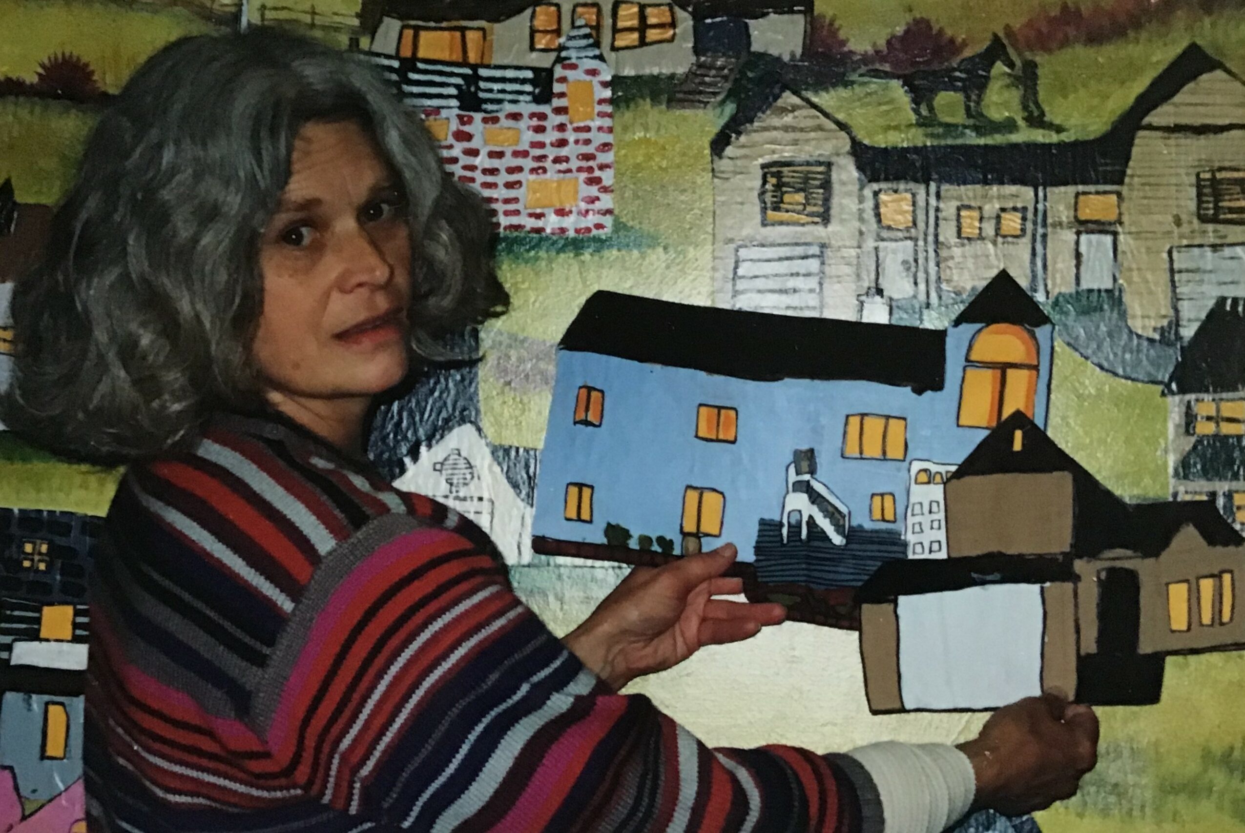 Woman with curly hair holding a painting in front of a colorful mural of houses and buildings.