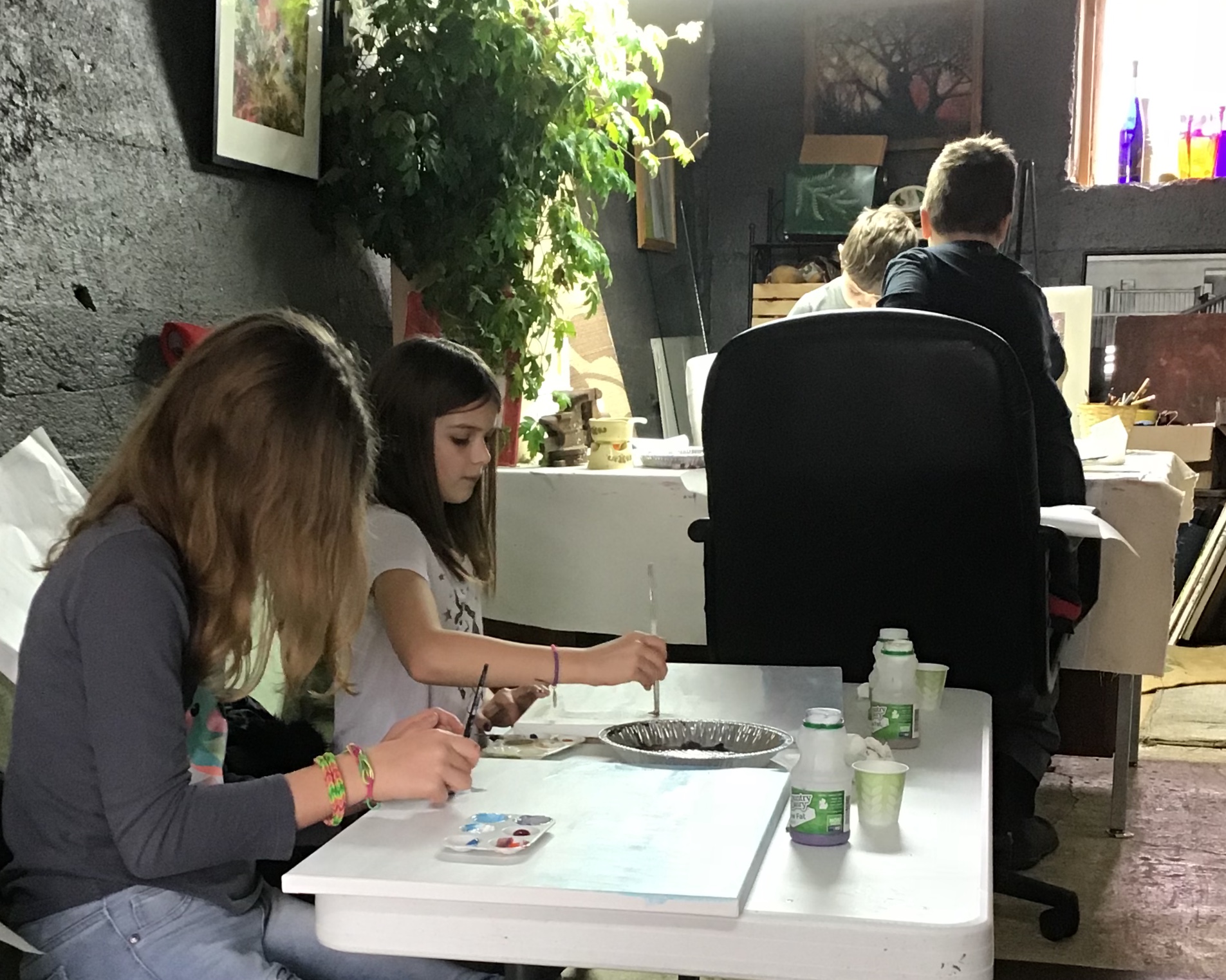 Two children sit at a table with art supplies, working on a project, in a room with plants and artwork.