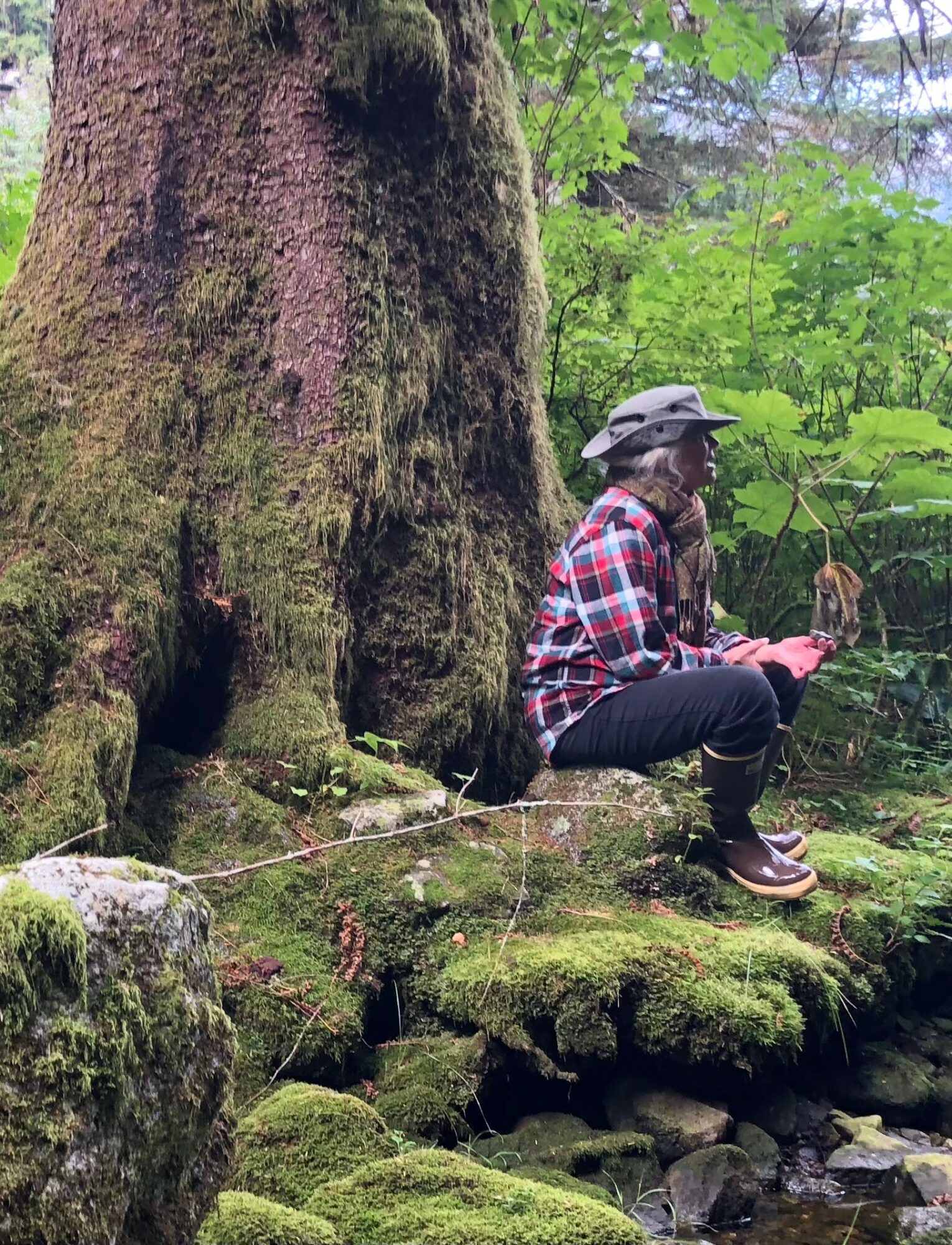 Person sitting on mossy log in forest, wearing plaid shirt, hat, and boots, surrounded by green trees and moss.