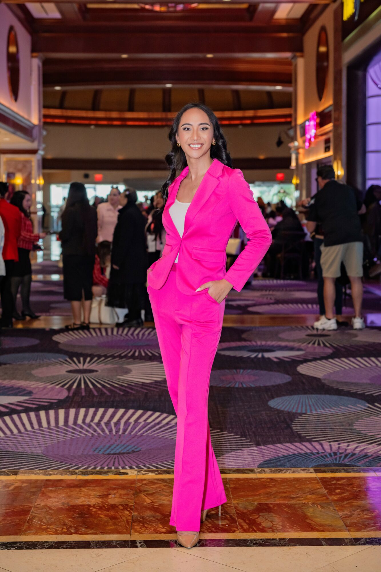 Woman in bright pink suit standing in a busy indoor venue with patterned carpet and people in the background.