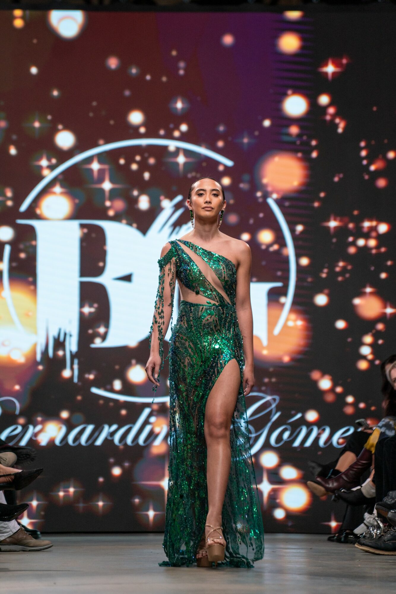 Model walking on runway in green sequined gown with thigh-high slit, background with sparkles and large letter B.