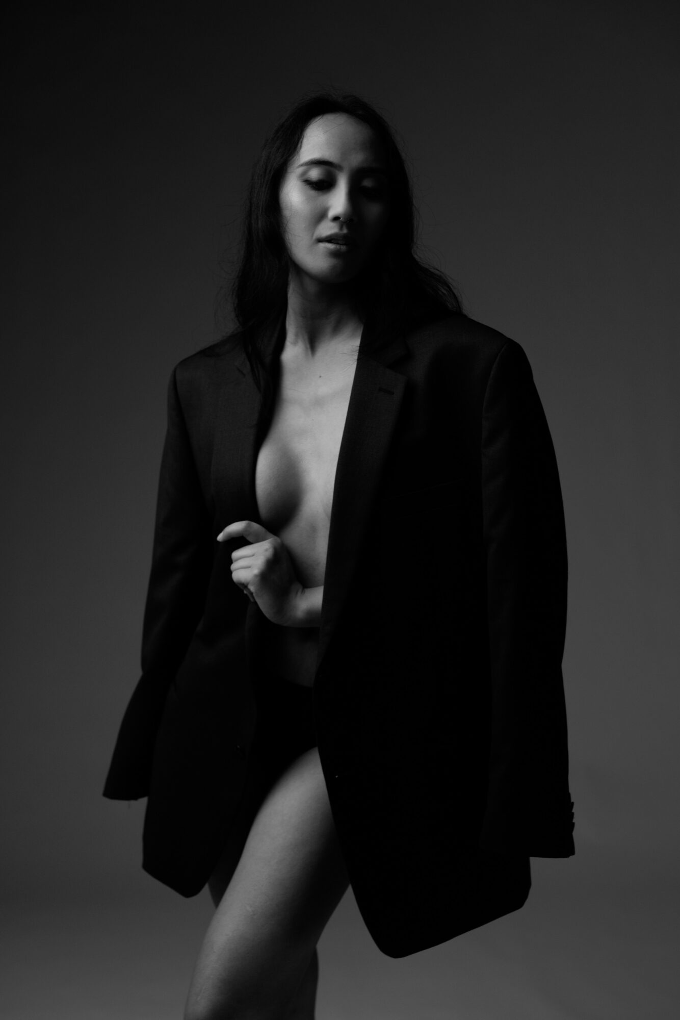 Woman with long hair wearing a blazer, partially nude, looking down, in black and white.