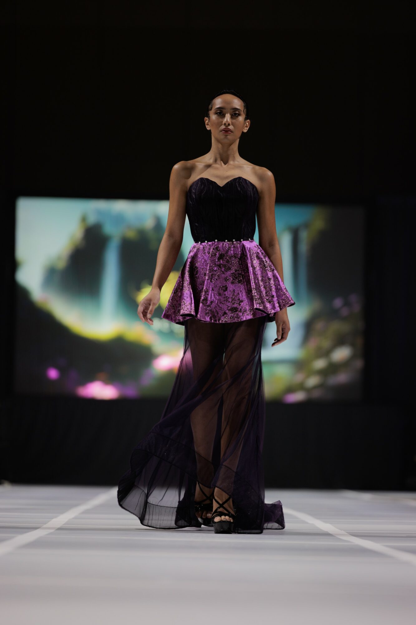 Fashion model walking on runway in purple and black dress with sheer black skirt, background displays colorful abstract image.