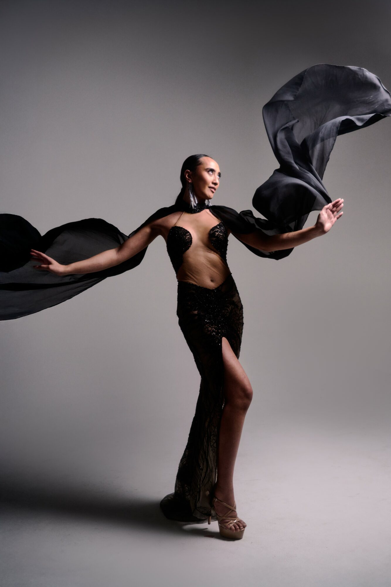 A woman in a black outfit holds flowing fabric, standing against a plain background, with arms extended and one leg slightly bent.