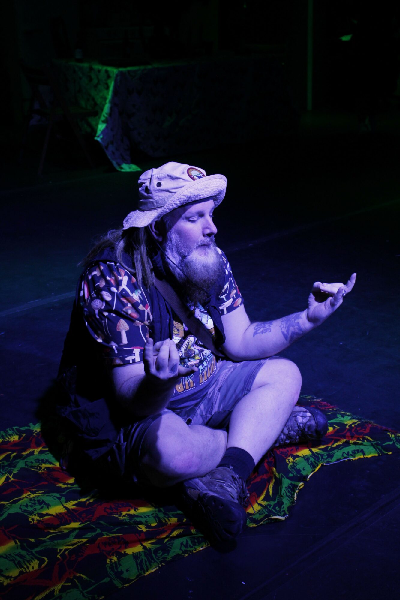 Two people lying on a colorful blanket in a dark room, one with a tattooed arm, both with eyes closed.
