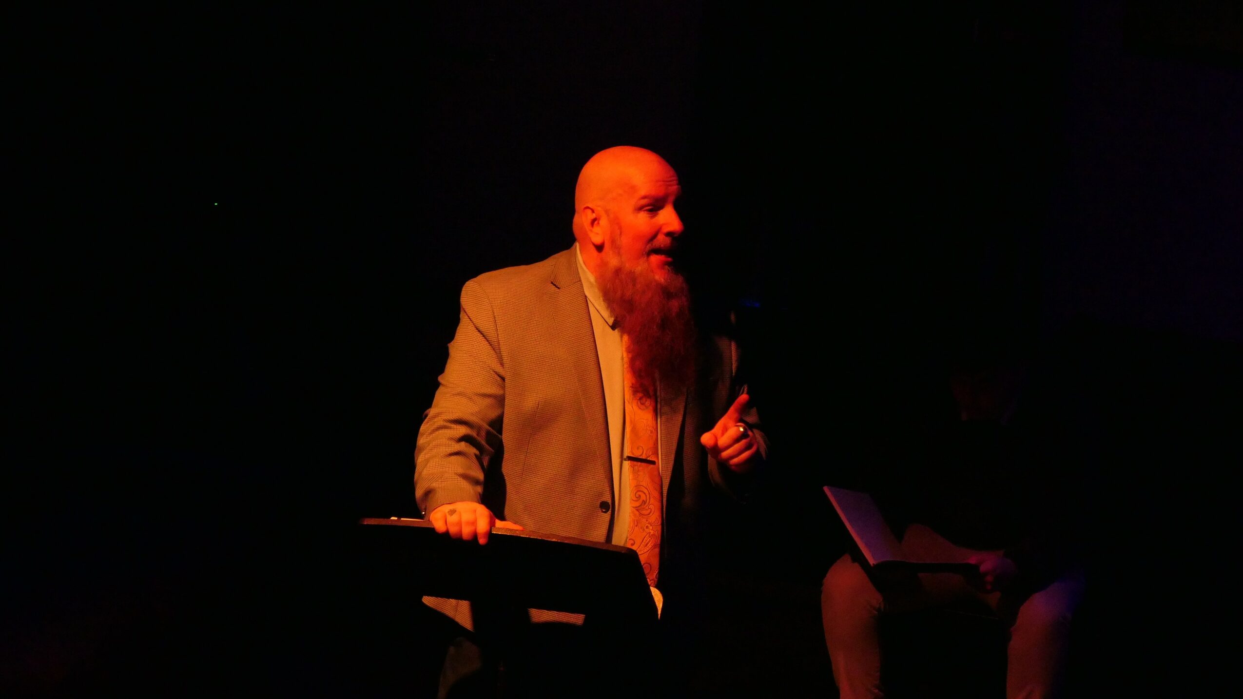 A bearded man in a suit speaking into a microphone on stage, with a dark background.