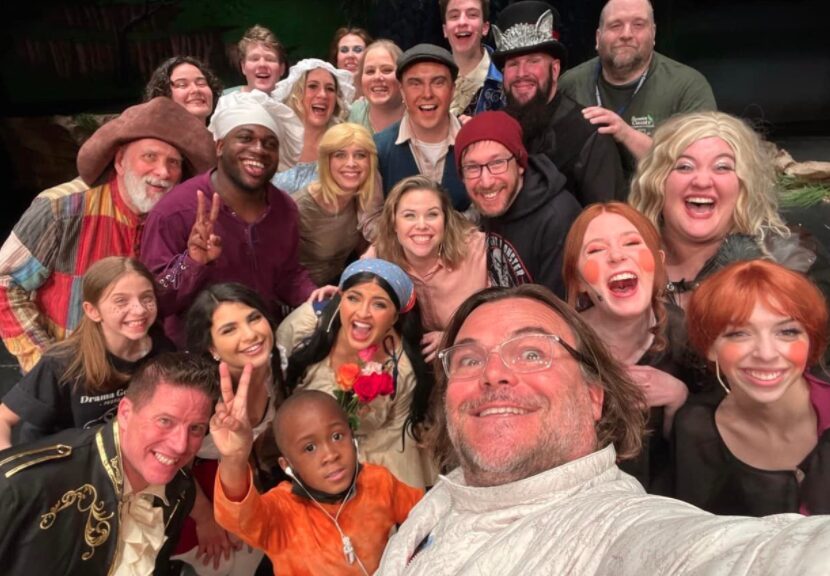 Group of diverse people smiling and posing for a photo indoors, some making peace signs, with a dark background.