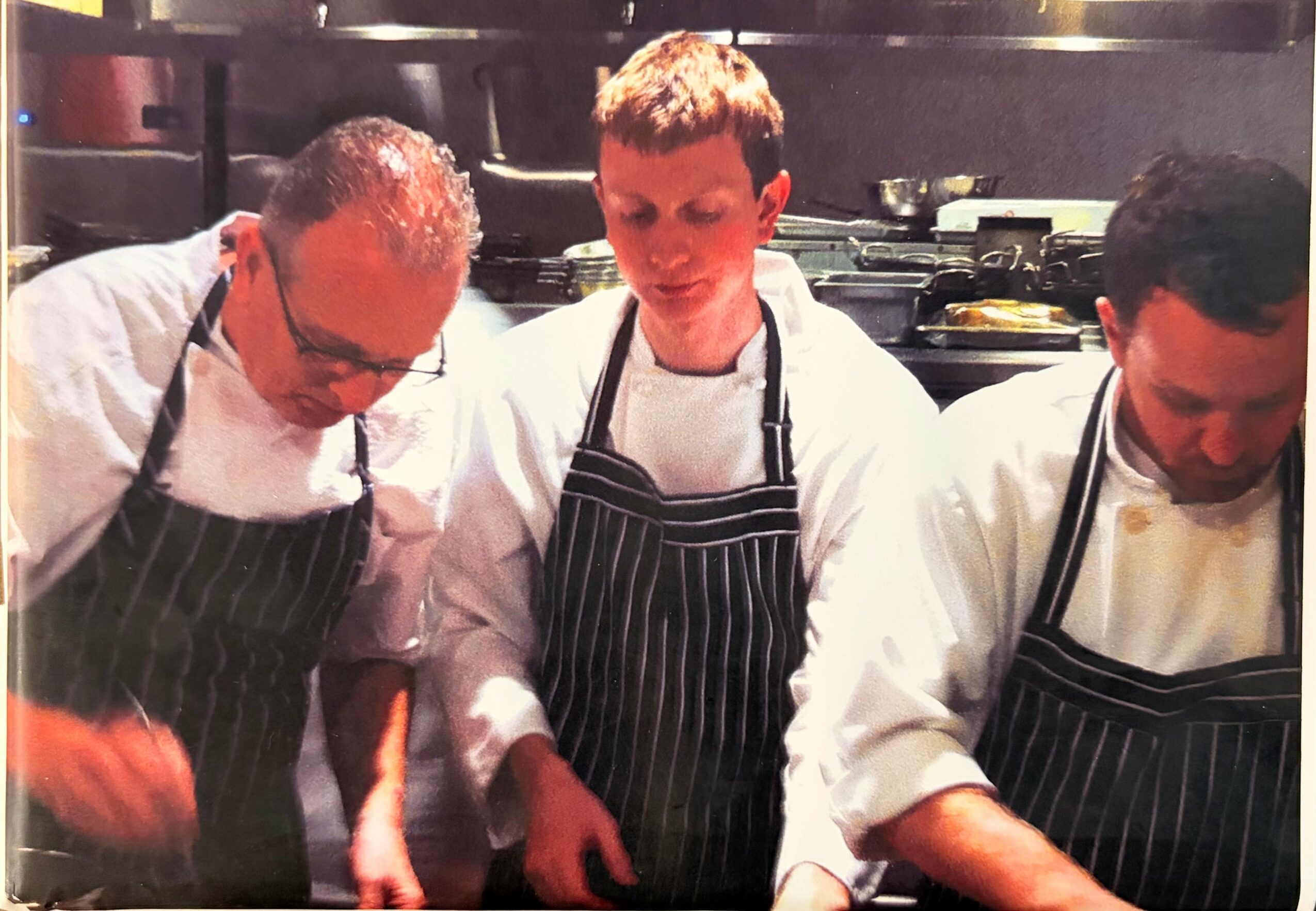 Three chefs in white uniforms and striped aprons working in a kitchen, focused on their tasks.