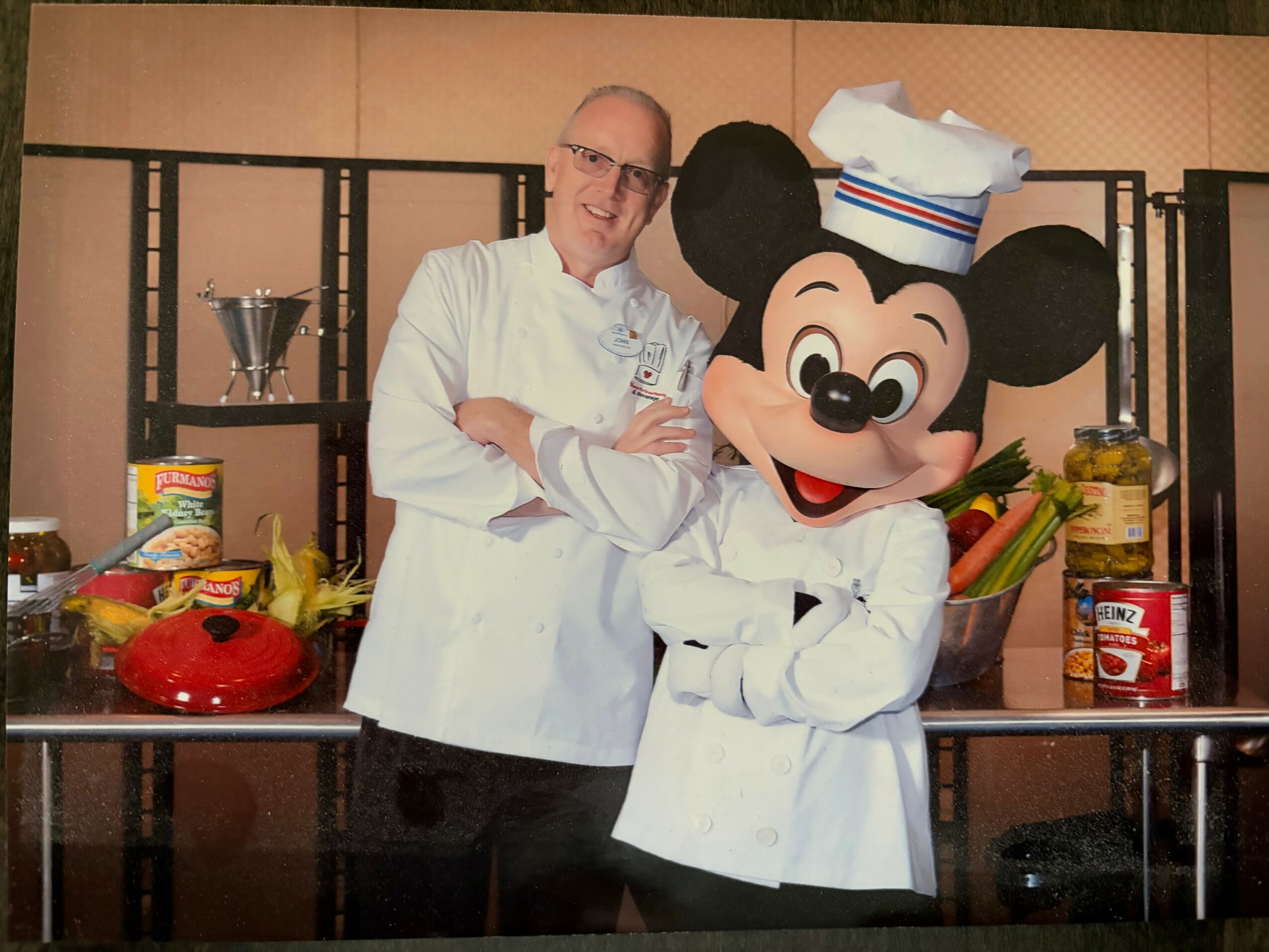Chef and Mickey Mouse in chef outfits behind a table with food and kitchen items.