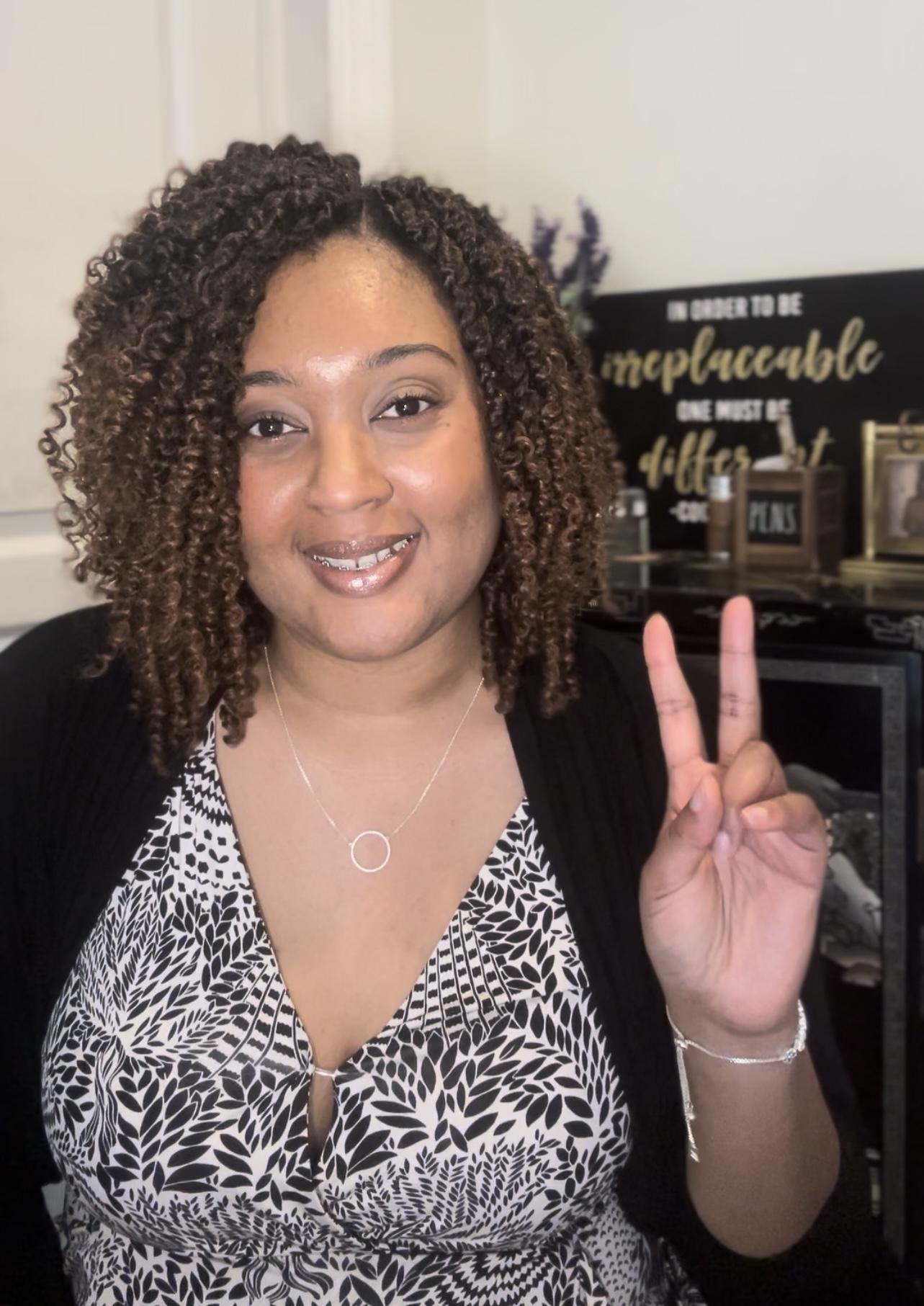 Woman with curly hair making a peace sign, smiling, in a room with decorative signs and items in the background.