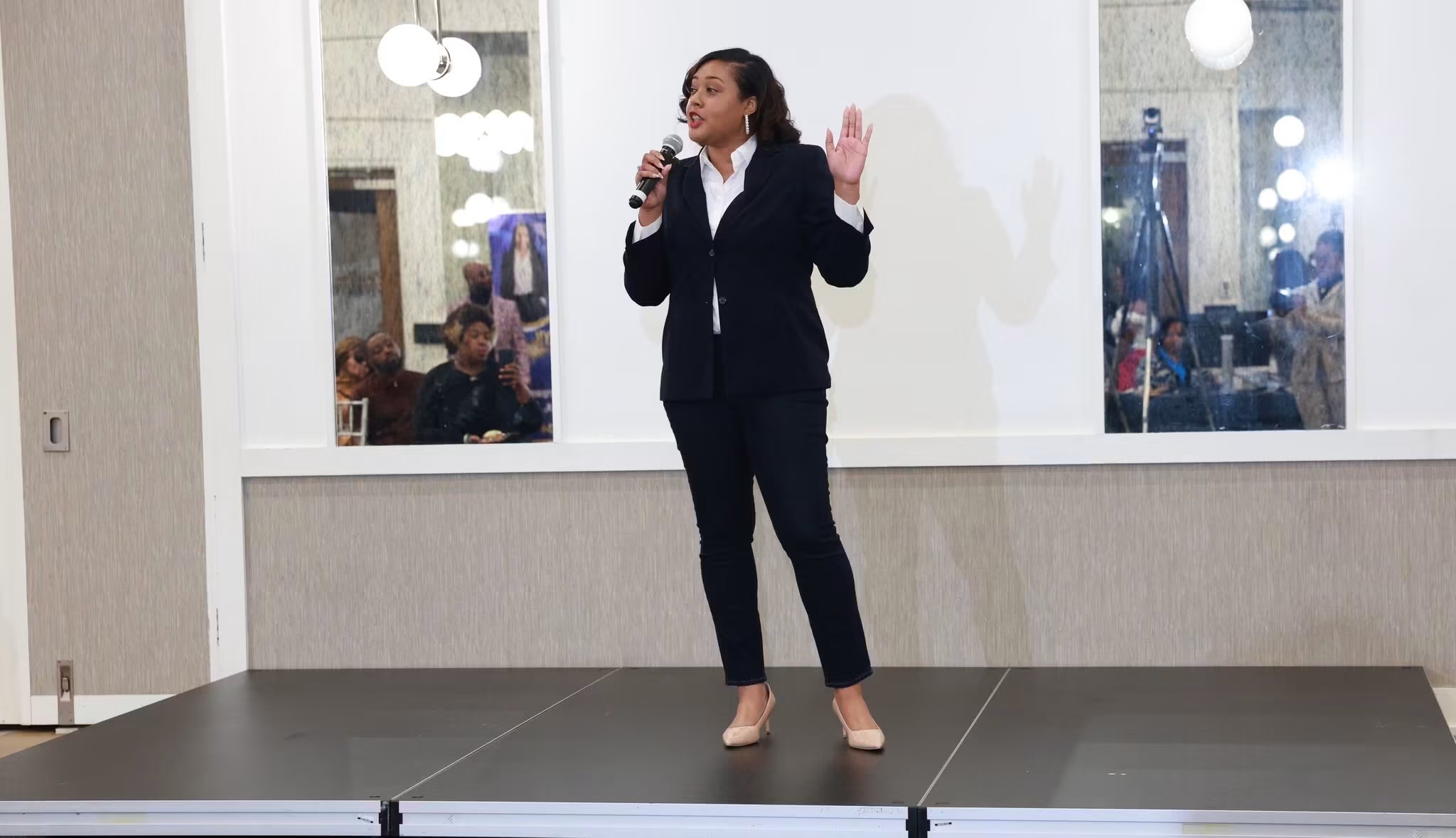 Woman in dark suit speaking into microphone on stage, with windows and audience in background.