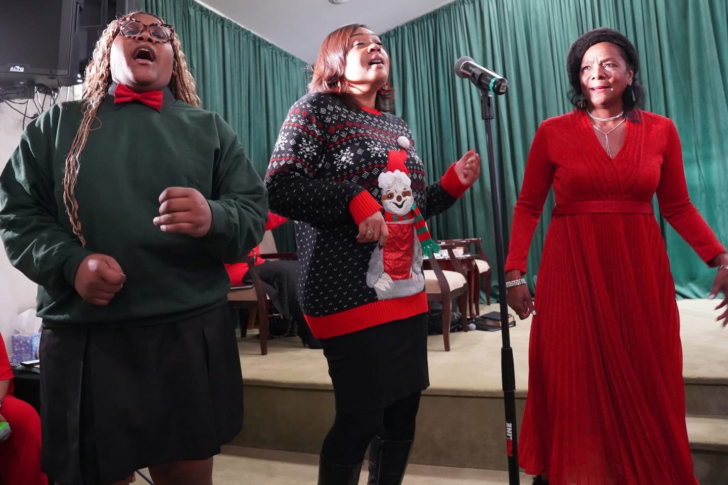 Three women singing on stage with a microphone, dressed in festive clothing, in front of a green curtain.
