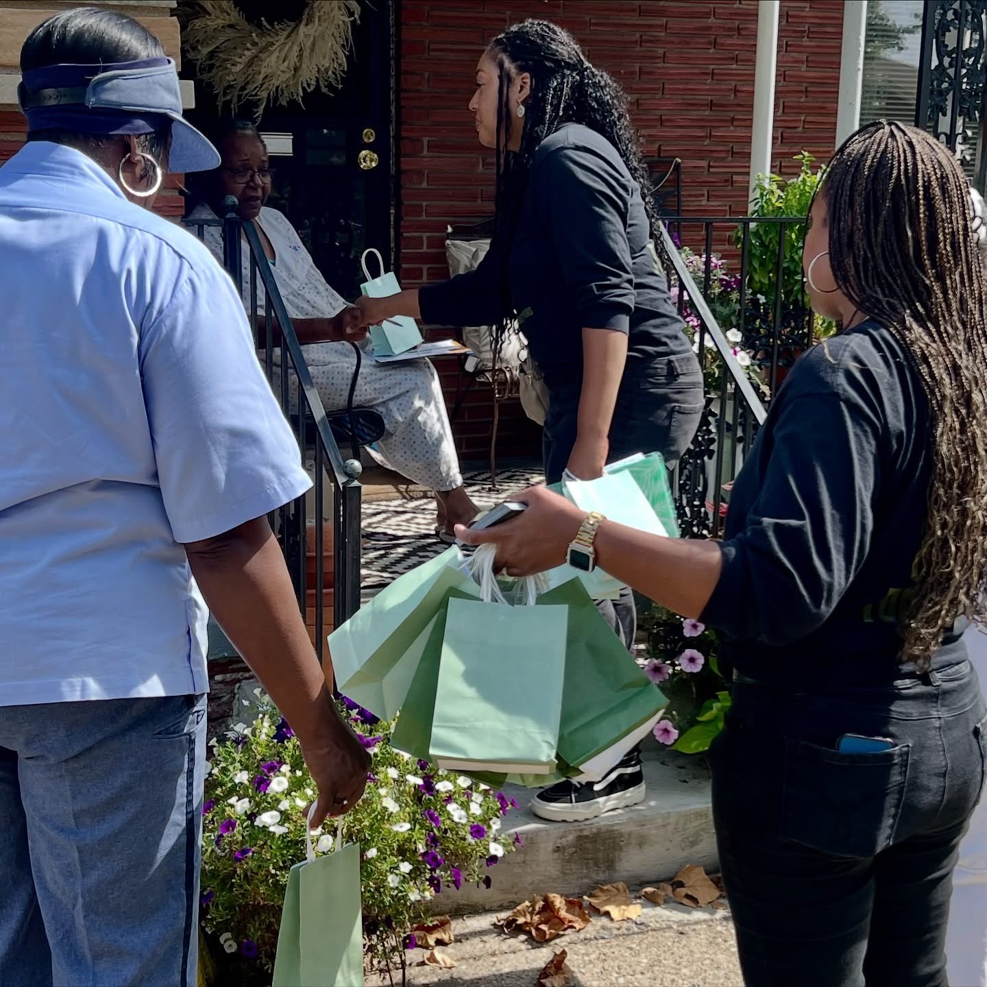 Group of people outside a building, some holding shopping bags, engaged in conversation, with flowers and greenery nearby.