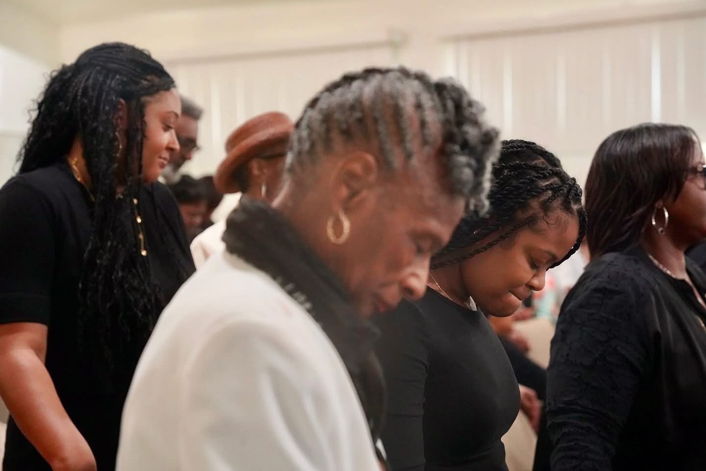 Group of people standing indoors, some with heads bowed, in a solemn or respectful moment.