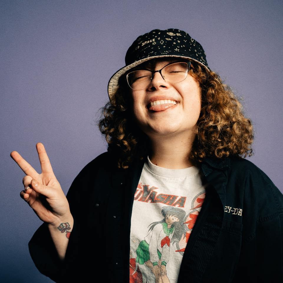 Person with curly hair, glasses, wearing a hat, smiling, making a peace sign, against a plain background.