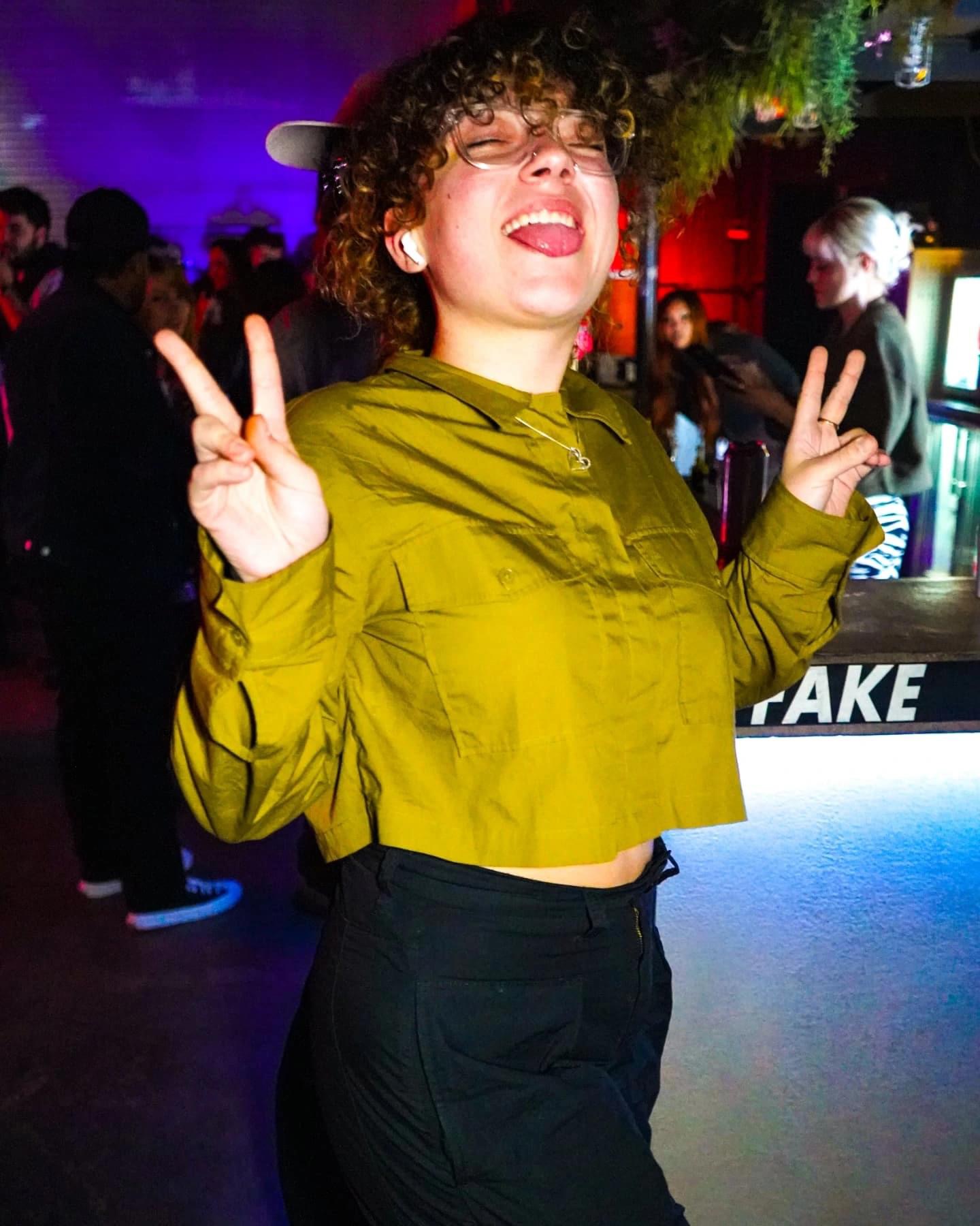 Young woman with curly hair smiling and making peace signs with both hands in a lively indoor setting.