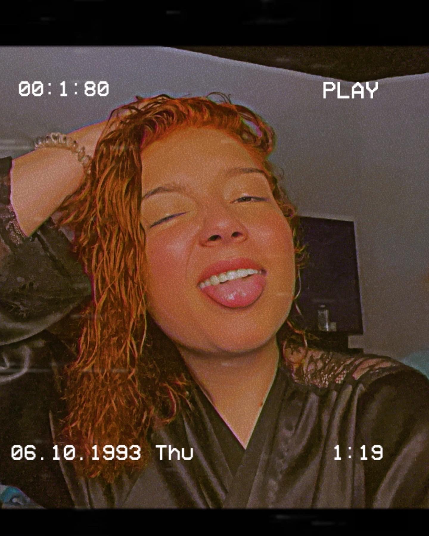 Young woman with curly hair sticking out her tongue, winking, in a room with a TV in the background.