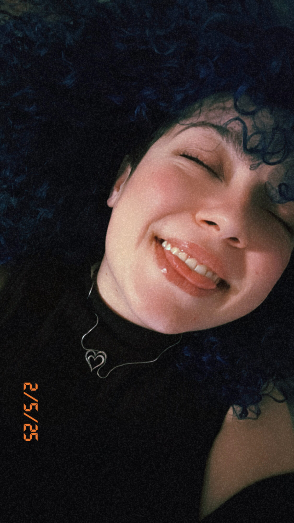 Person with curly hair smiling, eyes closed, wearing a necklace with a heart pendant, dark background.