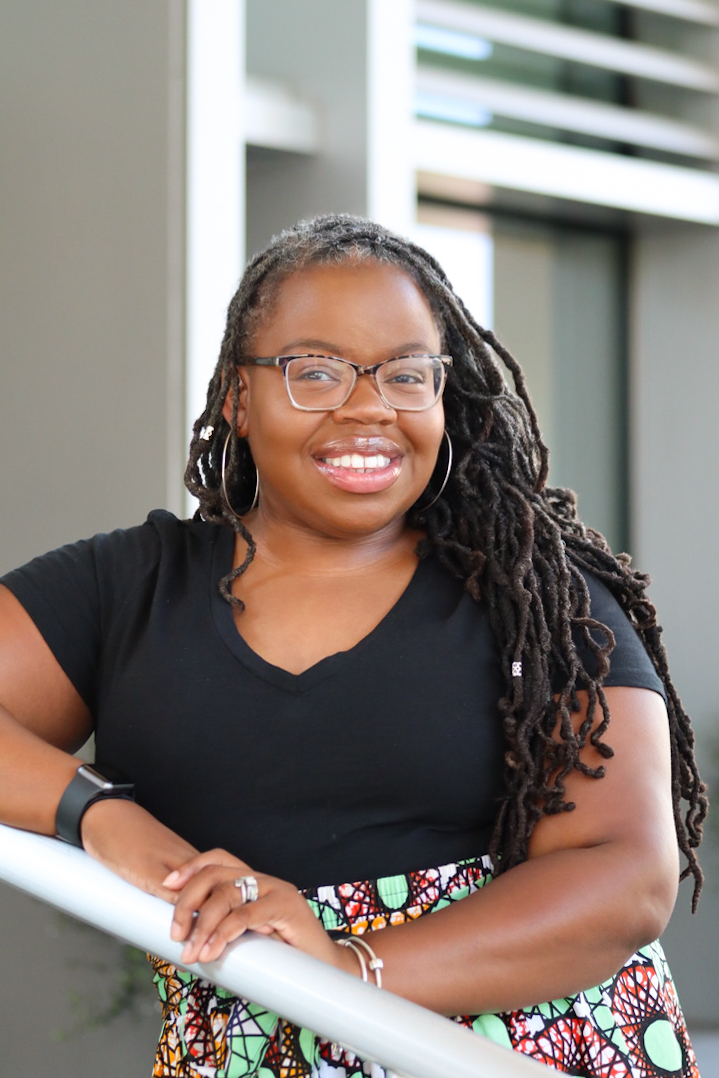 Smiling woman with glasses and long dreadlocks, wearing a black shirt, standing outdoors near a railing.