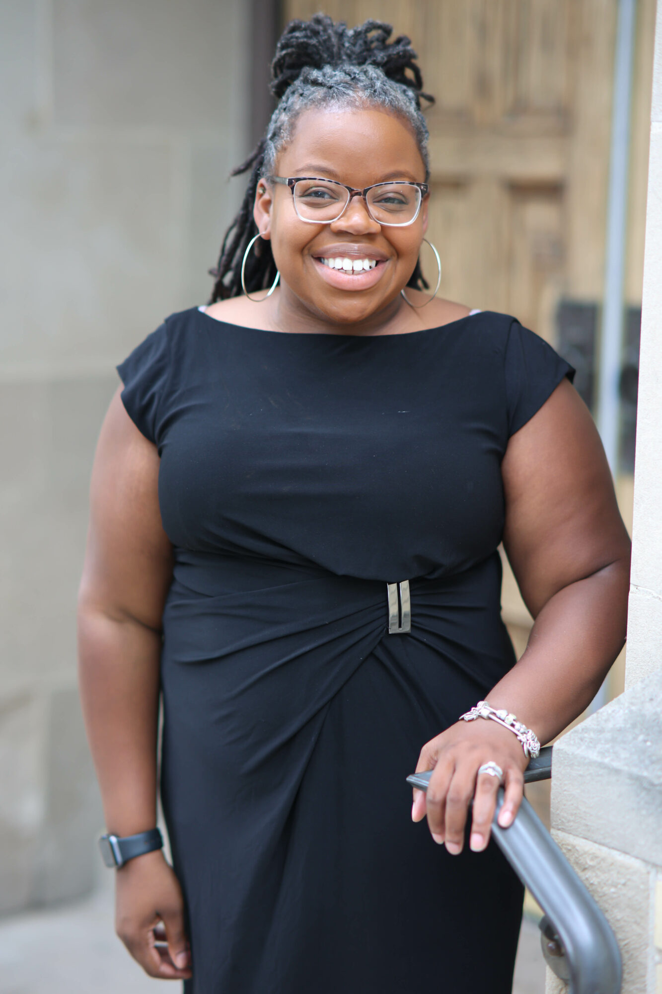 Smiling woman with glasses and braided hair in a black dress outdoors, holding a railing.