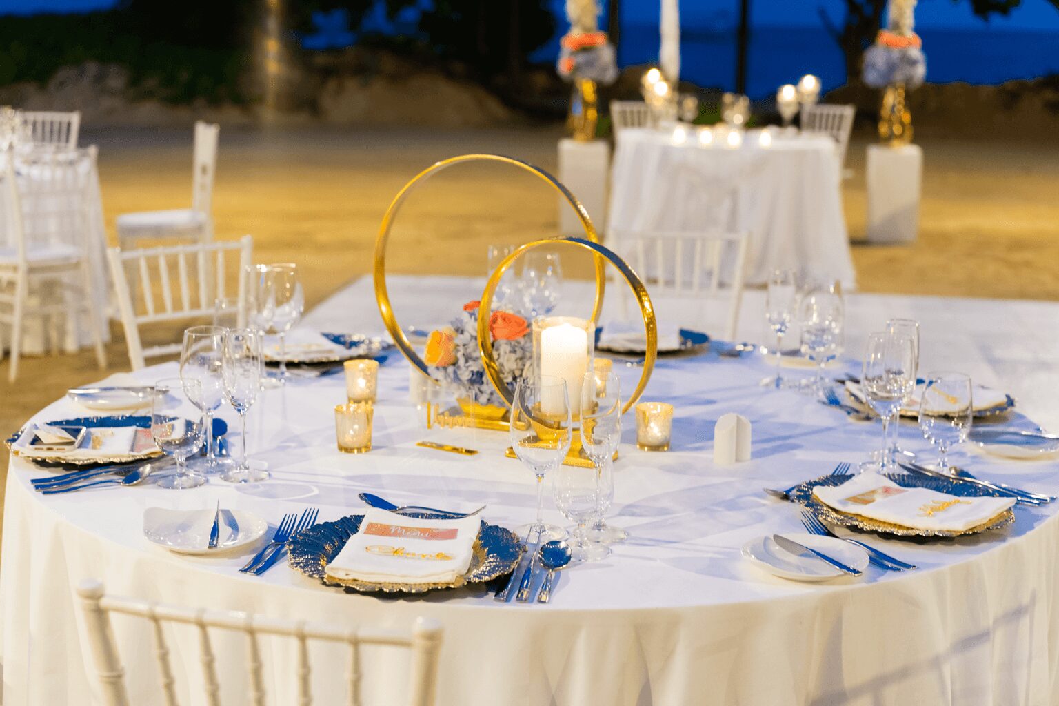 Round table set for a formal event with white chairs, floral centerpieces, and tableware, outdoors at night.