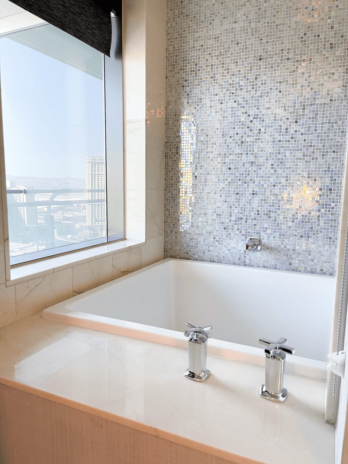Corner of a bathroom with a bathtub, mosaic wall, and large window showing a city view.