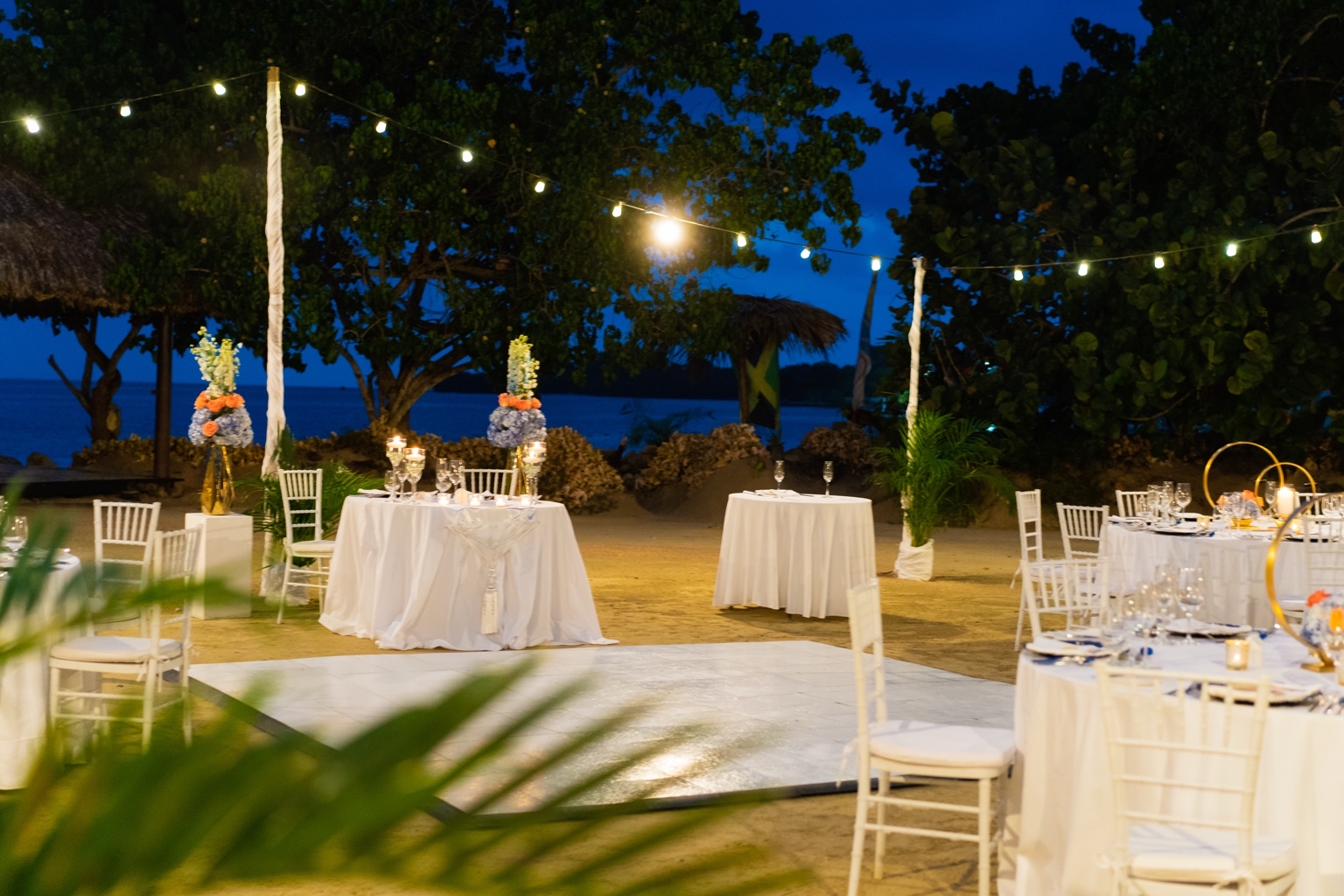 Outdoor event setup with tables, chairs, string lights, and trees at night.