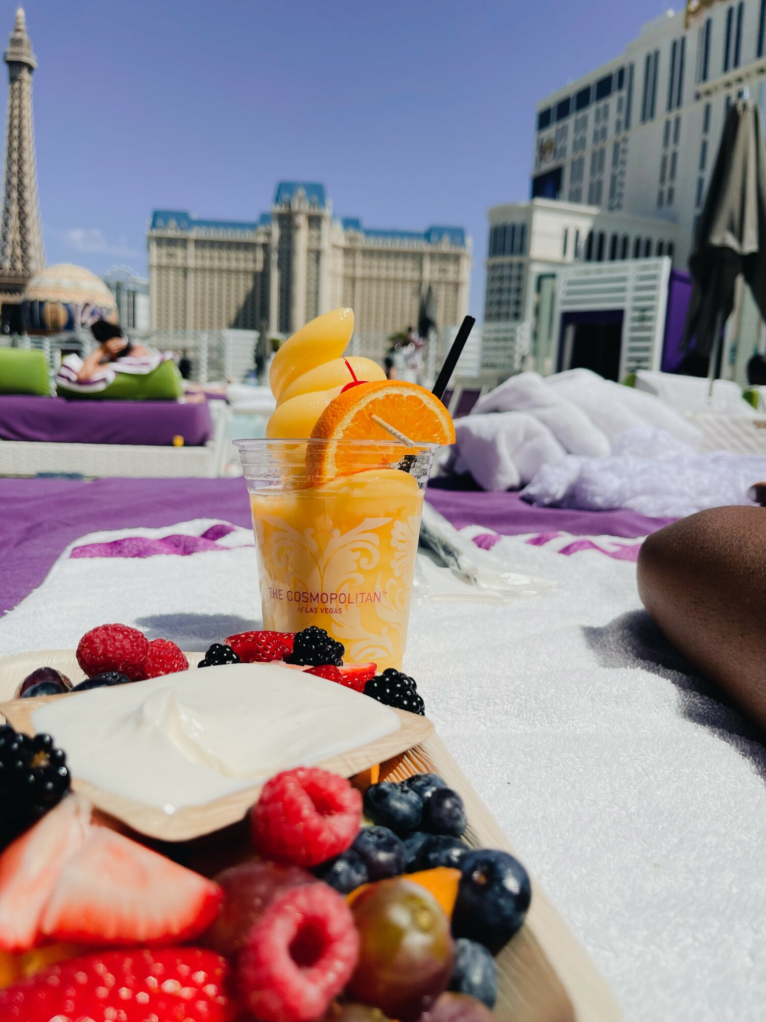 A rooftop scene with a drink, fresh berries, and a city skyline in the background, sunny weather, and lounge chairs.