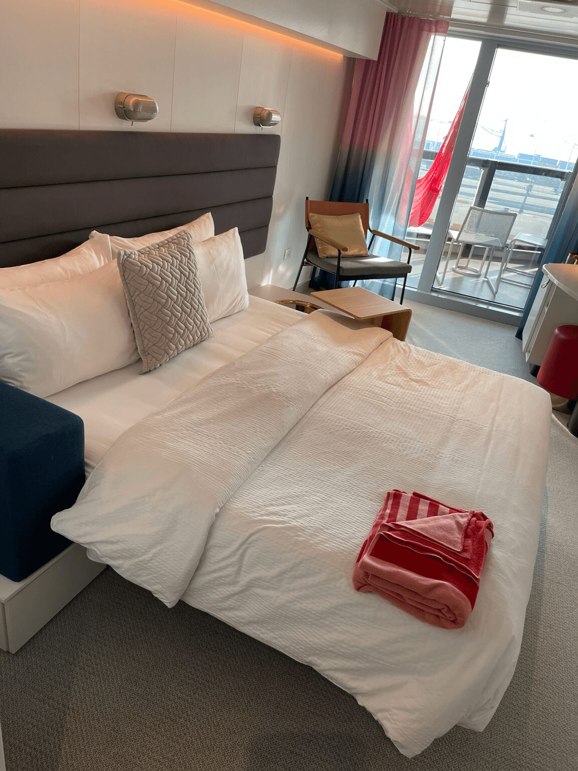 Unmade bed with white bedding, a pillow, a folded red towel, and a small table near a balcony with a view.