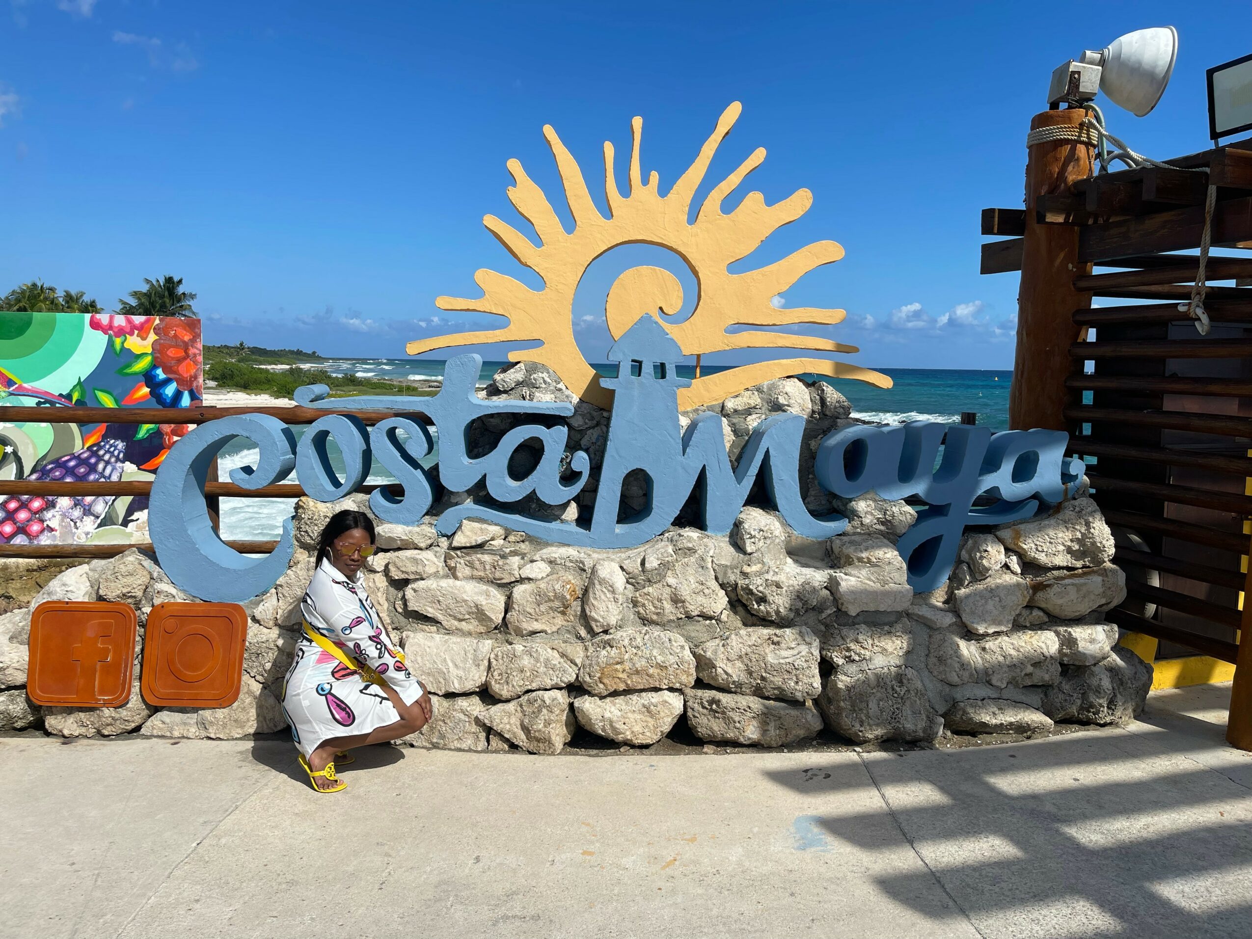 Person crouching near a sign that reads 'Costa Maya' with a sun and wave design, against a blue sky and ocean background.