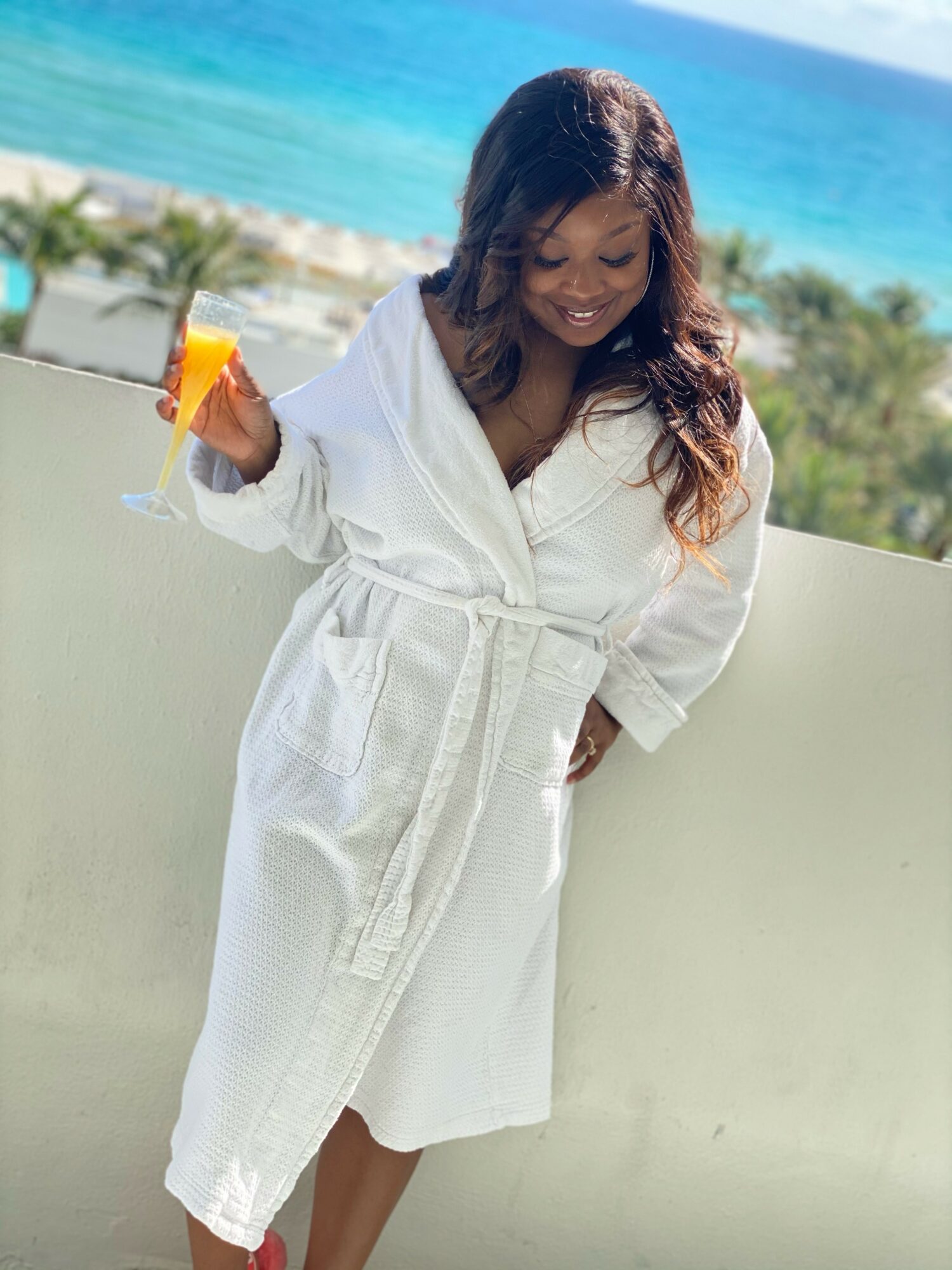 Woman in a white bathrobe holding a drink with an orange slice, standing outdoors near the ocean with palm trees.