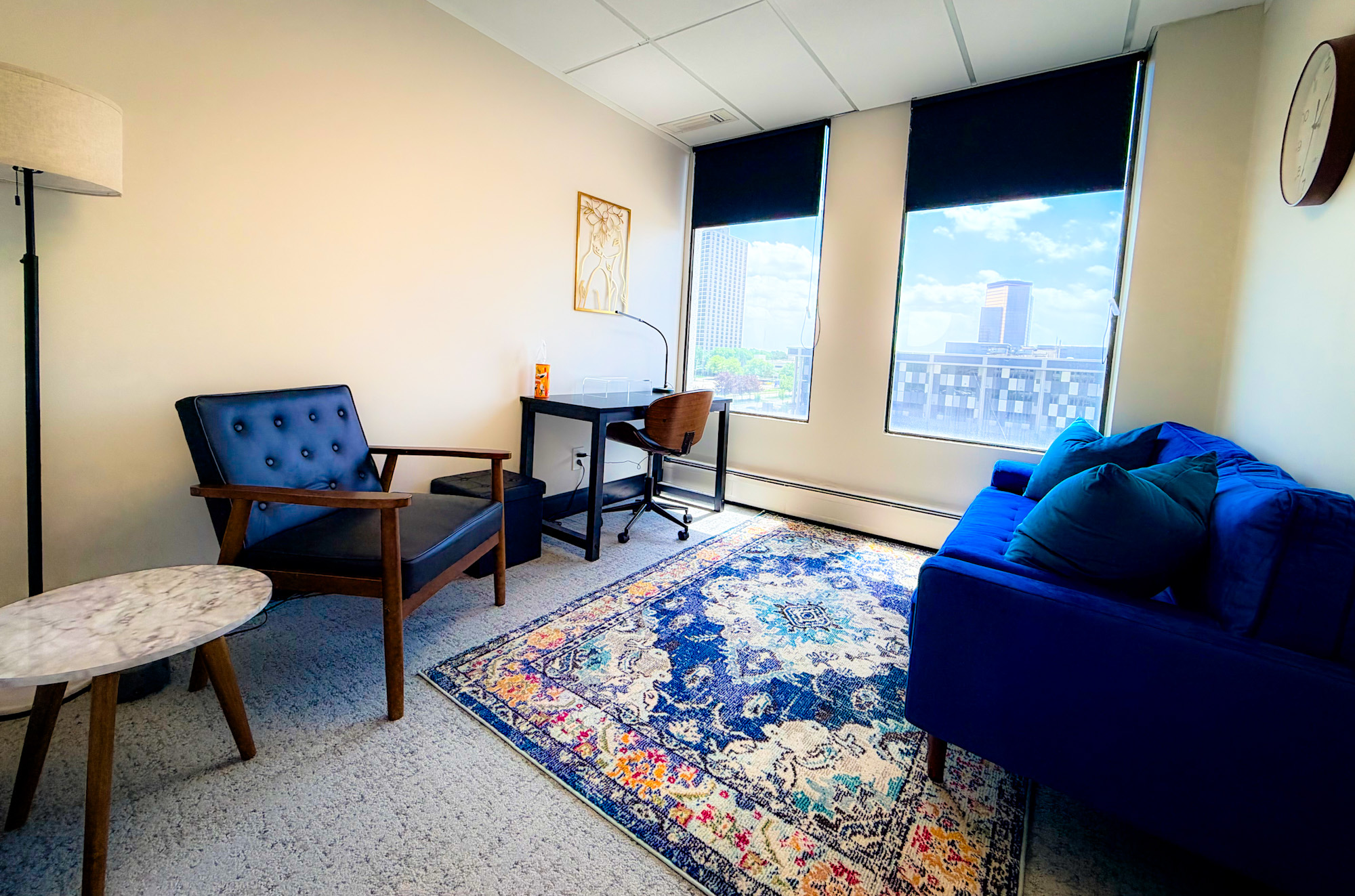 Office room with blue sofa, chairs, desk, windows, and a colorful rug, bright with natural light.