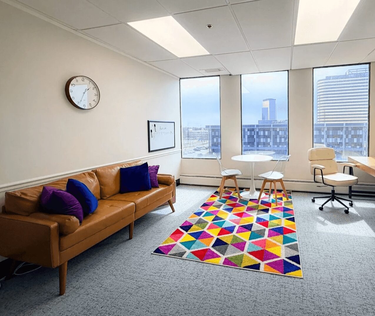 Office room with a brown sofa, colorful geometric rug, white chairs, large windows, and a wall clock.
