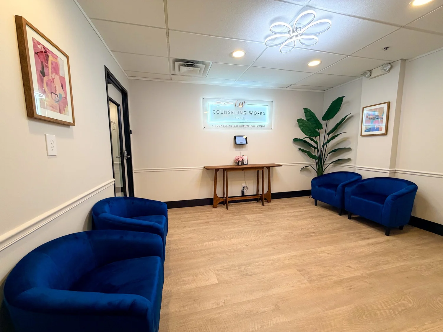 Waiting room with two blue sofas, a wooden table, a potted plant, and framed artwork on white walls.