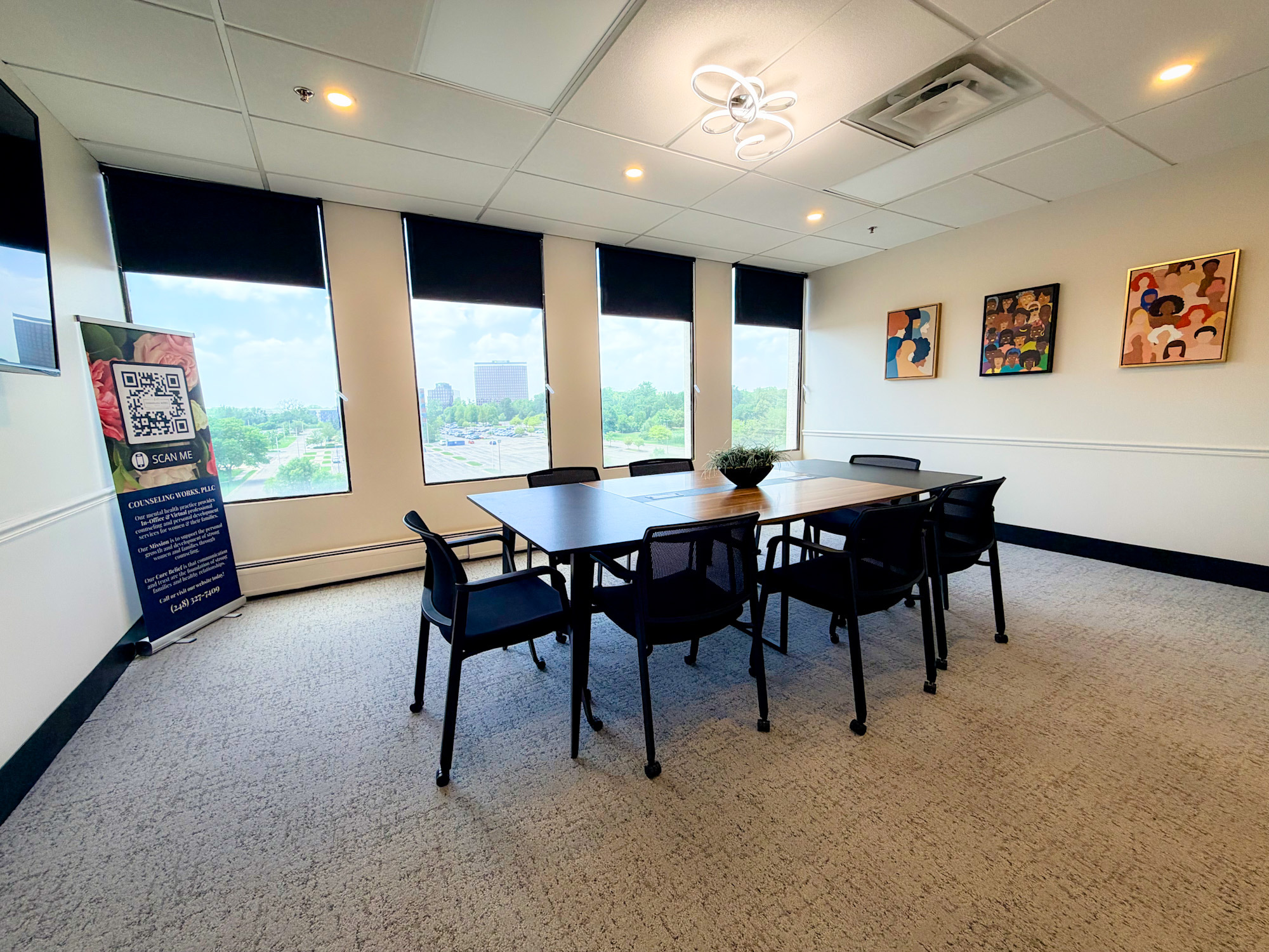 Conference room with six chairs around a table, large windows with city view, artwork on wall, carpeted floor.