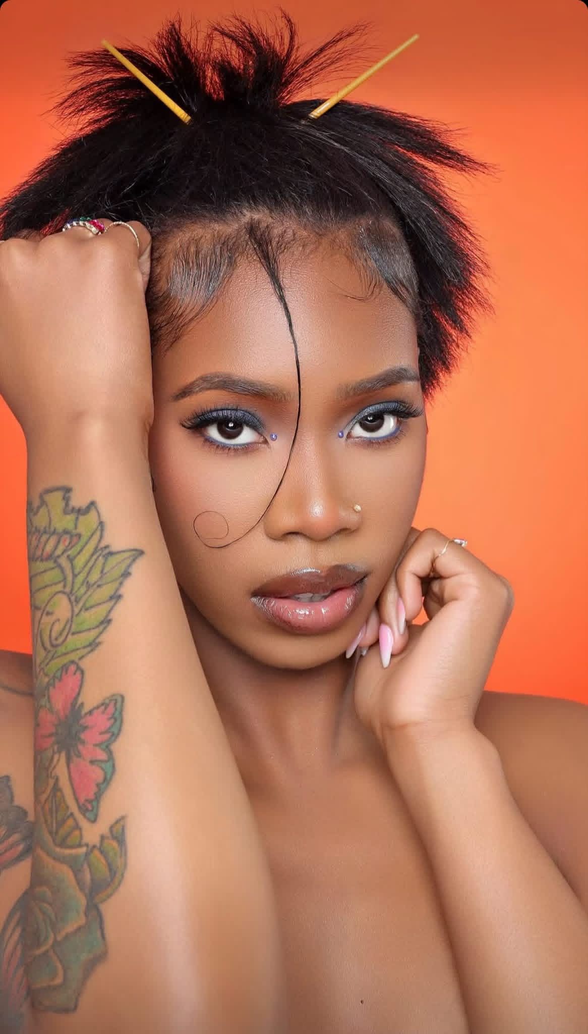 Young woman with short, spiky hair and tattoos on her arm, posing against an orange background.