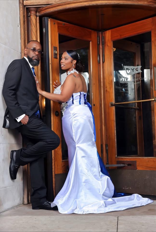 Man in a suit and woman in a white gown pose outside a wooden door, holding hands and looking at each other.