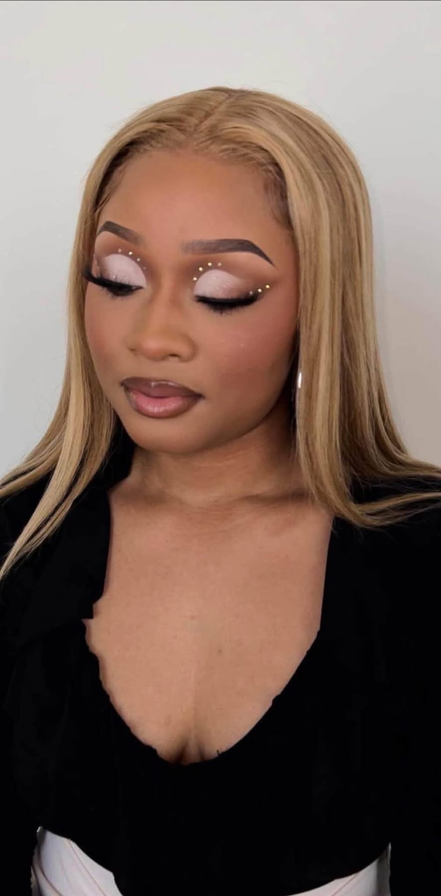 Woman with blonde hair and makeup, eyes closed, wearing a black top, against a plain background.
