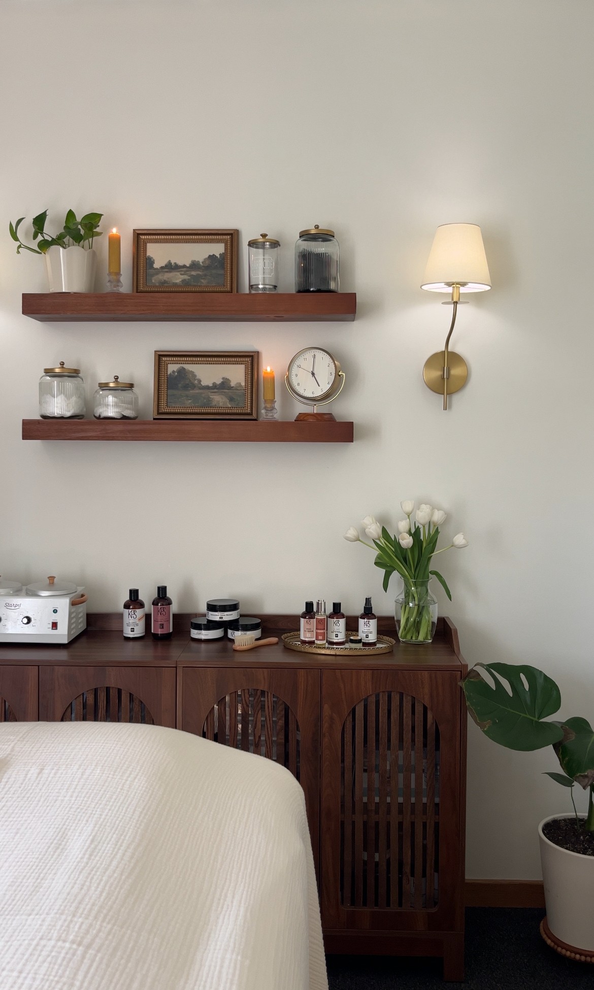 Wall with two wooden shelves holding framed pictures, jars, and a clock, with a wall-mounted lamp and a potted plant nearby.