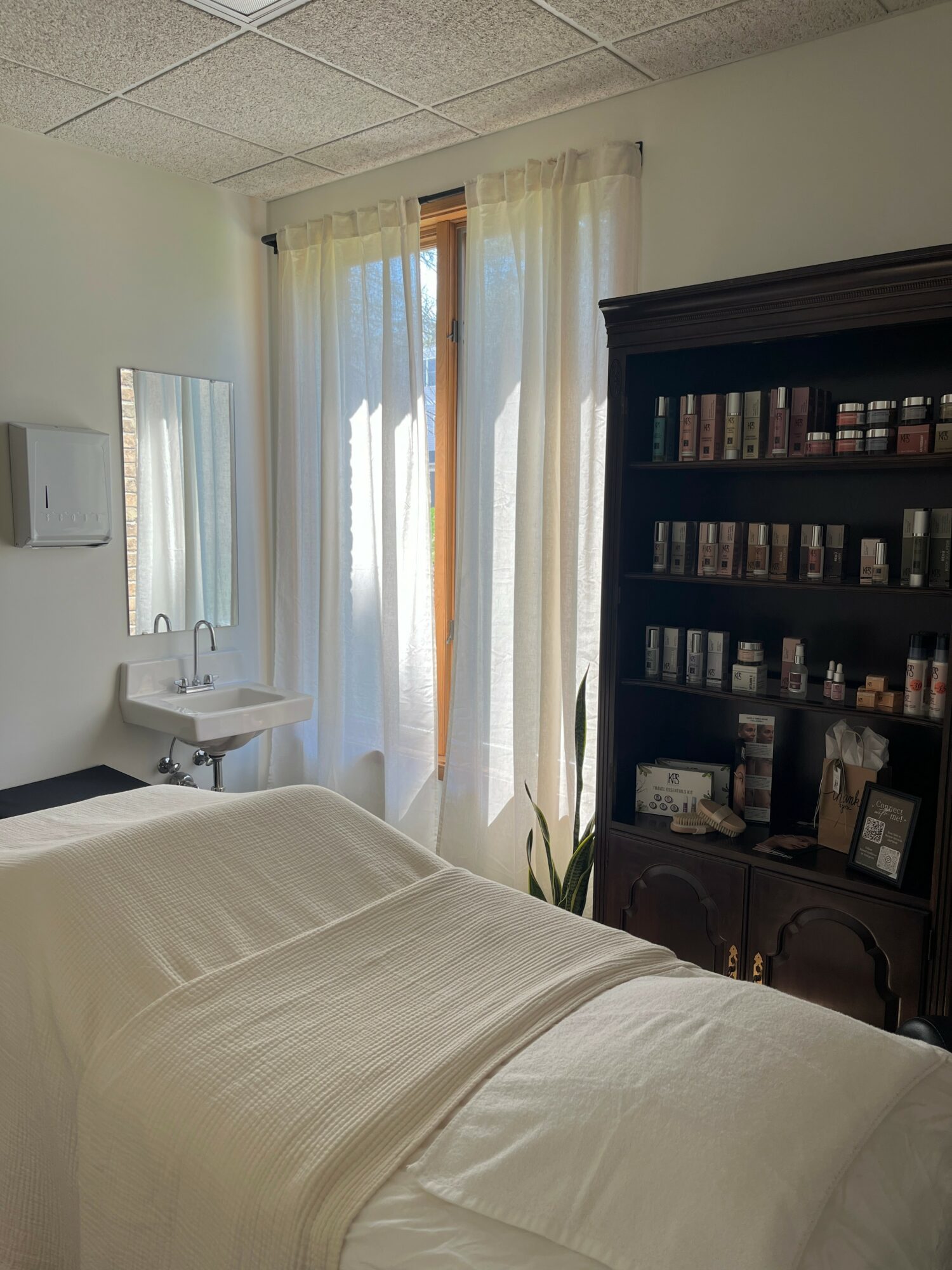 A bedroom with a bed, a window with curtains, a mirror, a sink, and a dark wooden bookshelf filled with books.