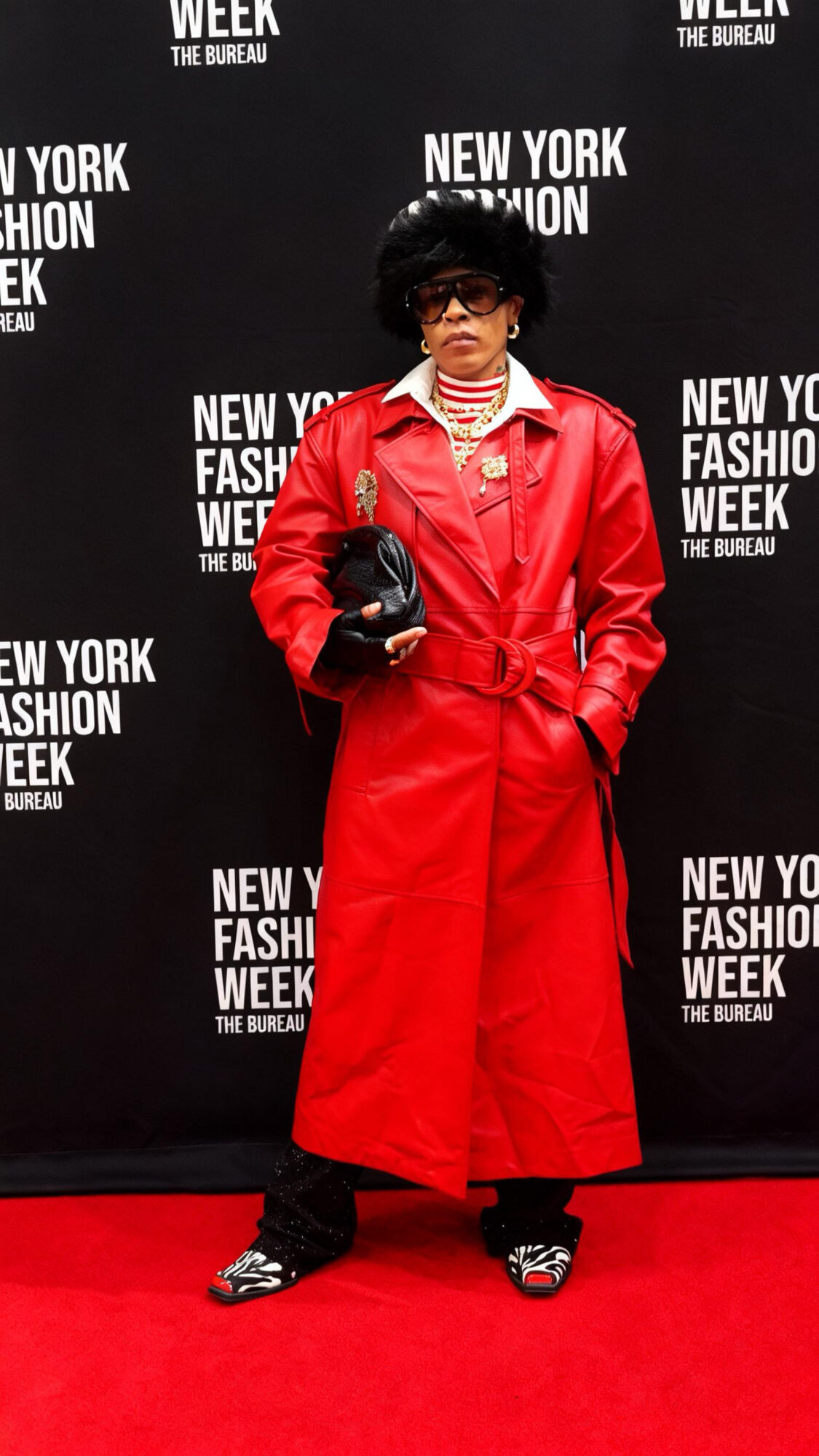 Person in red trench coat and sunglasses standing on red carpet at New York Fashion Week backdrop, holding a black bag.