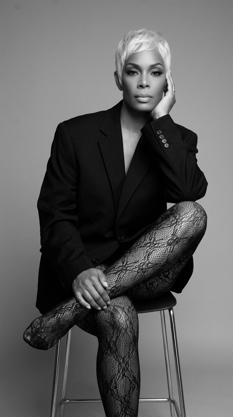 Woman with short platinum hair wearing a blazer and patterned tights, sitting on a stool with one hand on her face.