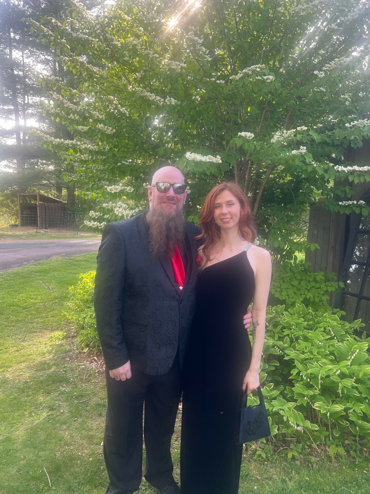 Two people standing outdoors in a garden with green trees and grass, one wearing a dark suit and the other in a black dress.