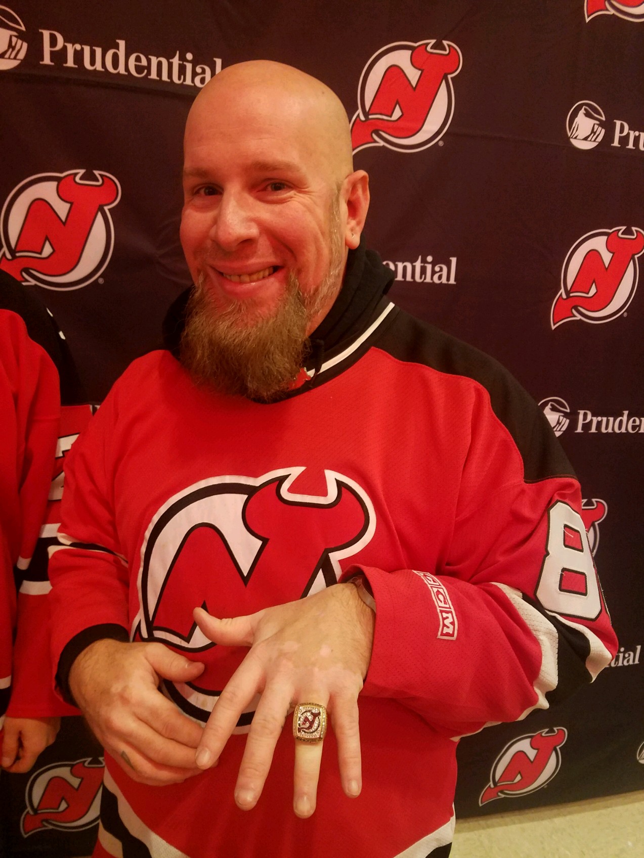 Man with a beard smiling, wearing a red New Jersey Devils hockey jersey, holding up his left hand with a ring, standing in front of a black background with team logos and