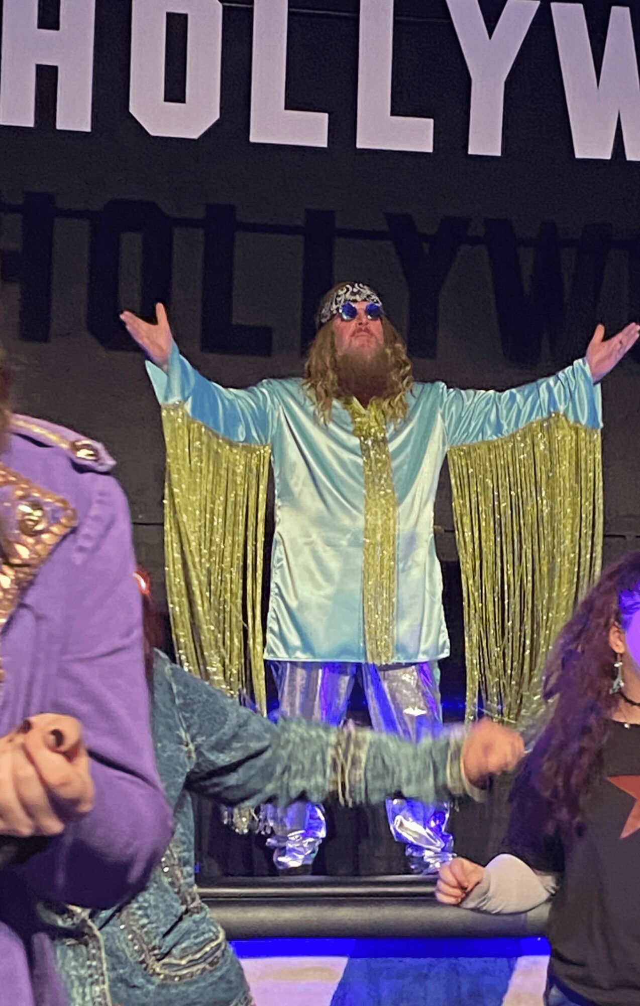 Person with long beard and sunglasses wearing a shiny, colorful costume with arms raised, stage background with 'HOLLYWOOD' text.