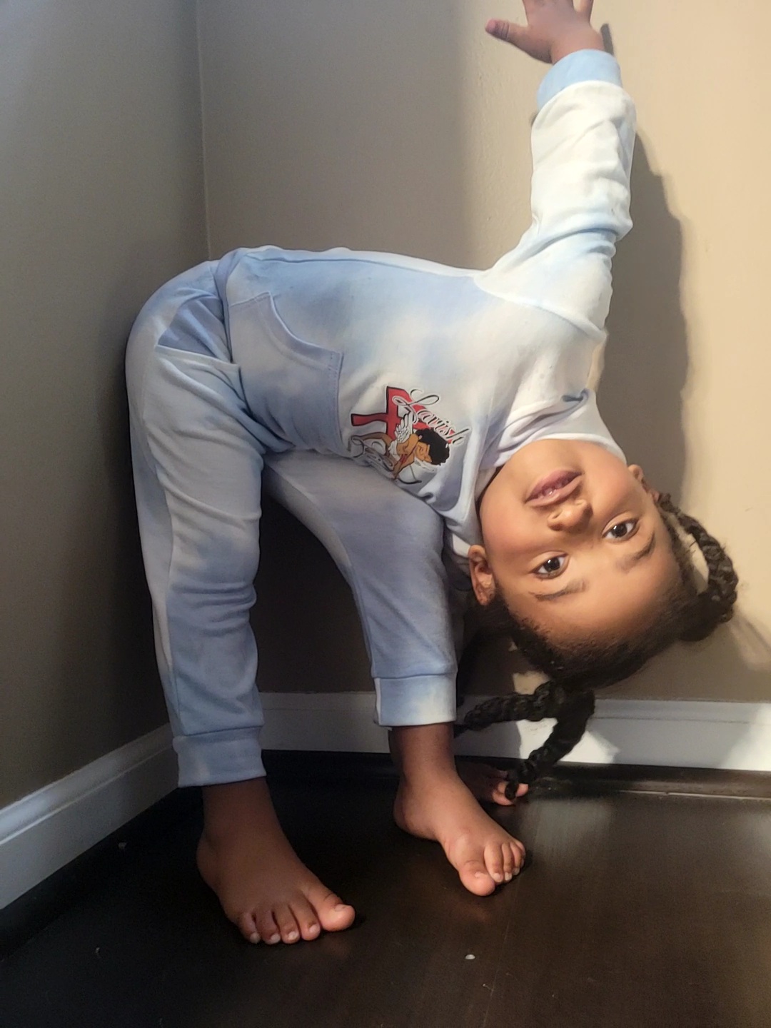 Child in pajamas doing a headstand against a corner wall, with one arm raised and face upside down.