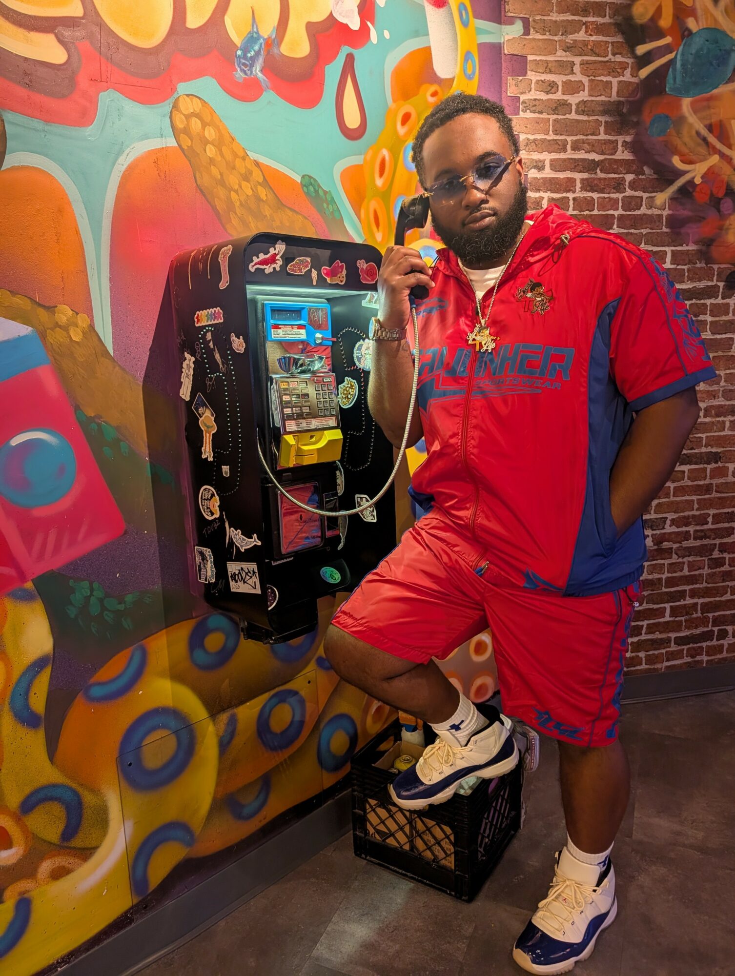 Man in red and blue sportswear with glasses, standing next to a colorful mural and a vintage payphone, holding the receiver.